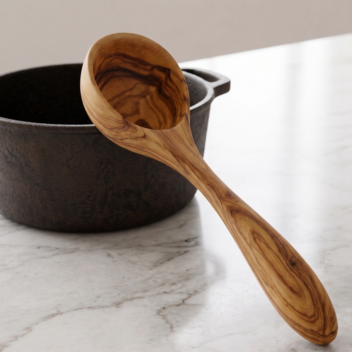 An olive wood ladle on the edge of a dark cast iron pot on a white marble countertop.