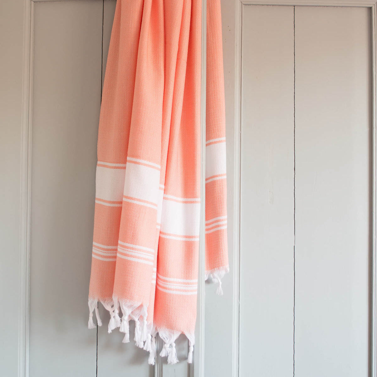 A hanging orange and white striped fouta towel with a fringed edge, displayed on a white door with a light gray background.