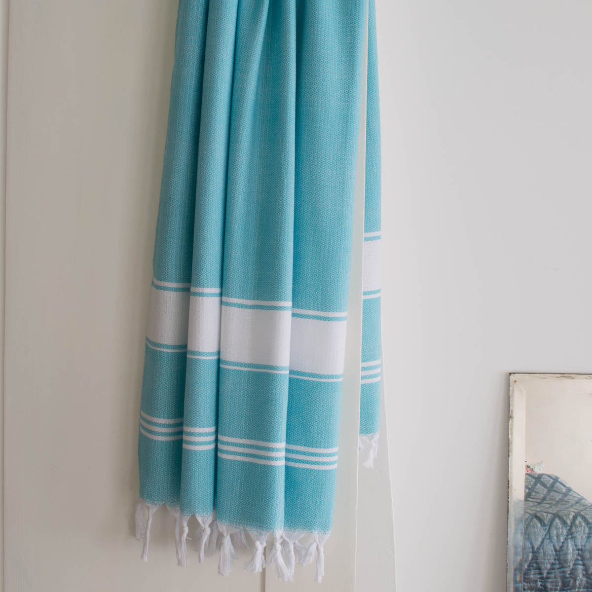 A turquoise blue Fouta towel with white stripes is hanging on a door, displaying its full width and fringed texture.