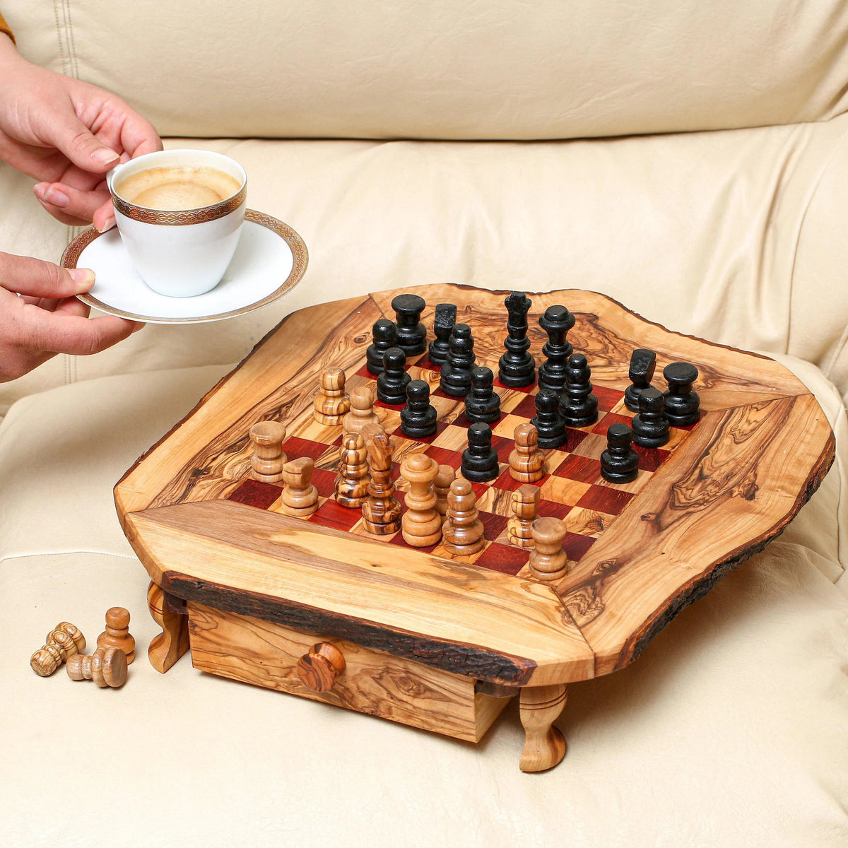 A person holding a cup of coffee over a handcrafted olive wood chess set with pieces arranged on the board, placed on a light-colored sofa.