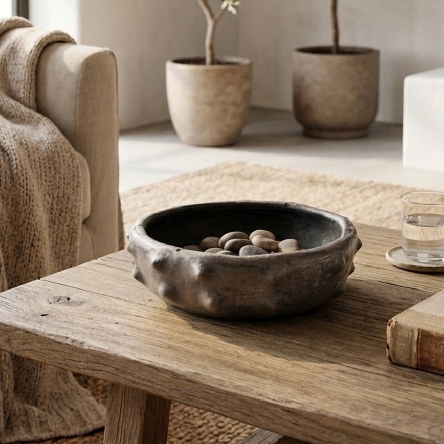 A rustic dark gray stone bowl filled with smooth river stones, styled on a weathered wooden coffee table in a cozy living room setting.
